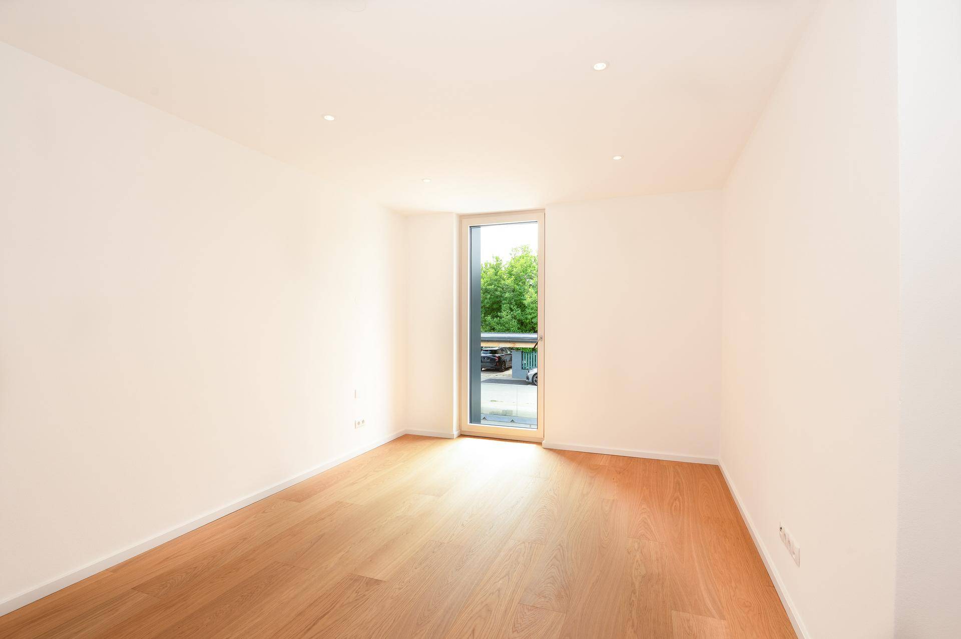 Leeres Zimmer mit hellem Eichenparkettboden und bodentiefem Fenster.