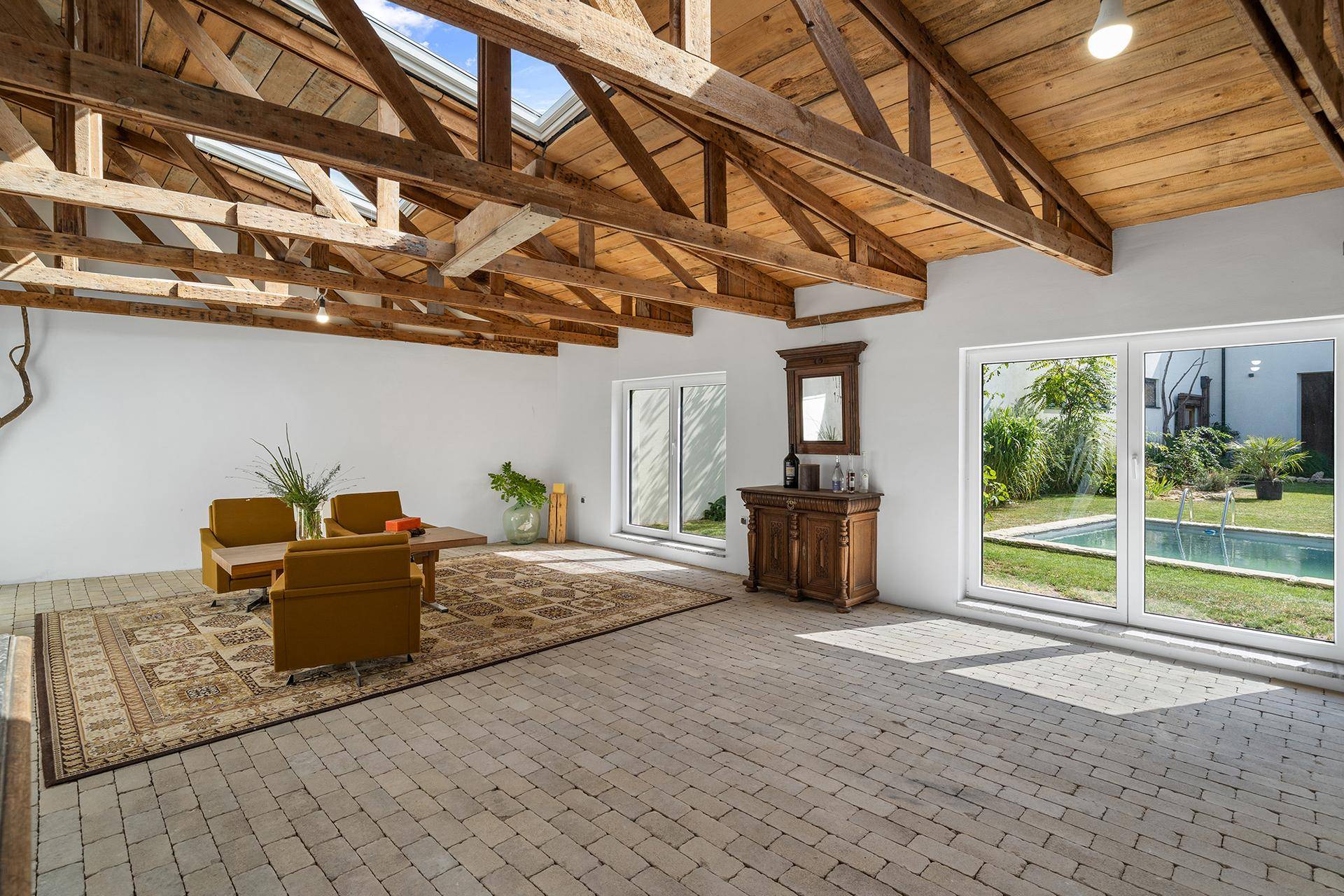 Großer Wohnbereich mit Holzbalken, Fliesenboden und Blick auf den Garten mit Pool.