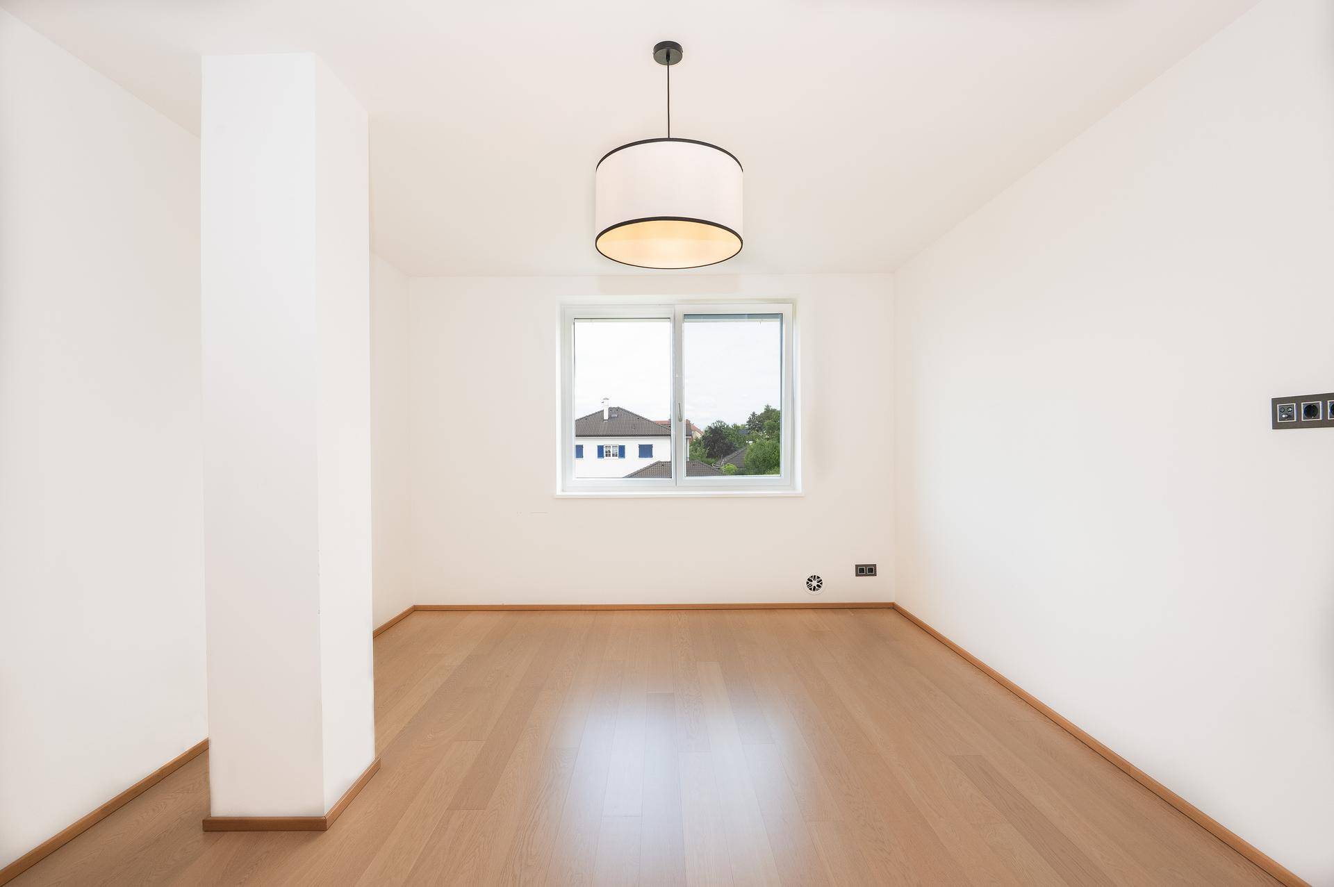 Heller, leerer Raum mit Holzboden, weißen Wänden und einem Fenster mit Blick ins Grüne.