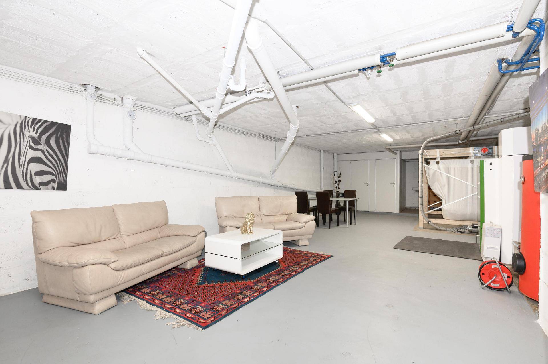 Ausgebauter Kellerraum mit Sitzbereich, Teppich und sichtbaren Versorgungsleitungen an der Decke.