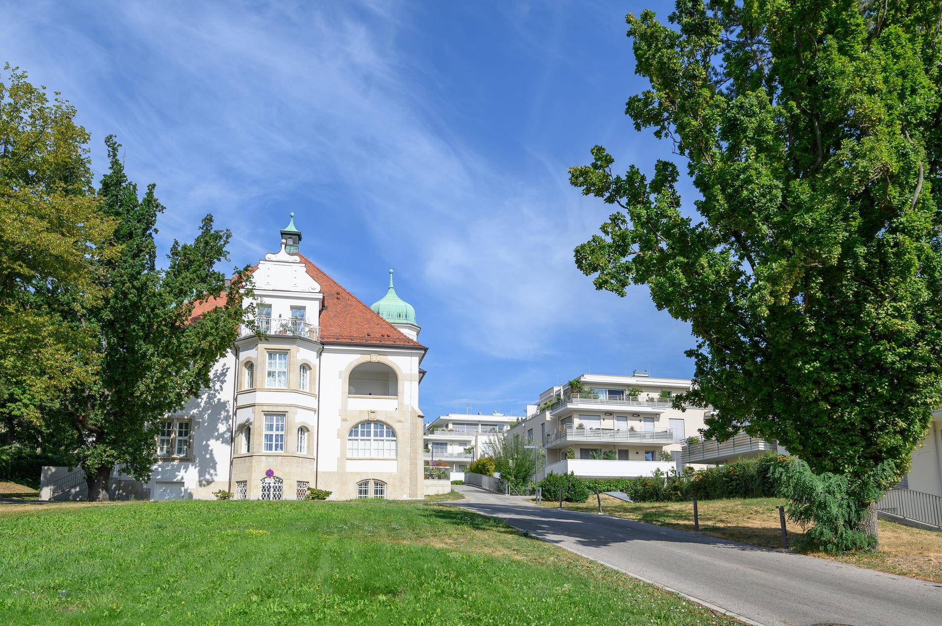 Historisches Gebäude mit markantem Dach und modernen Wohngebäuden im Hintergrund.