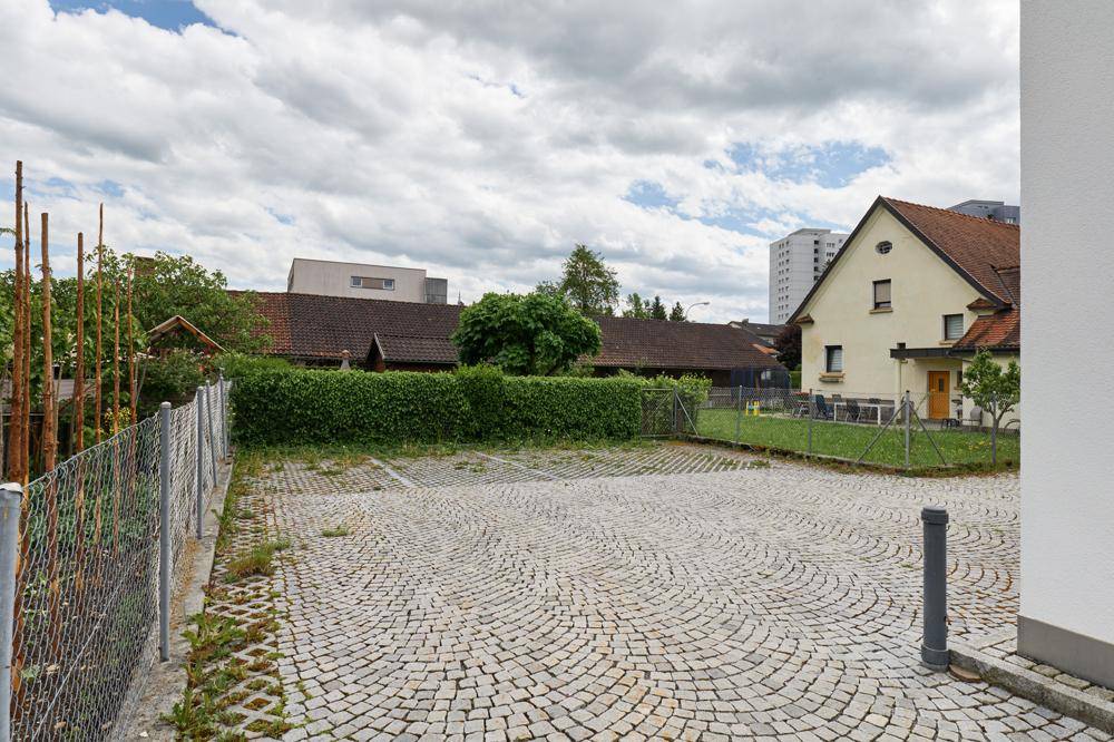 Außenbereich mit gepflasterten Parkplätzen und einer Hecke im Hintergrund unter bewölktem Himmel.