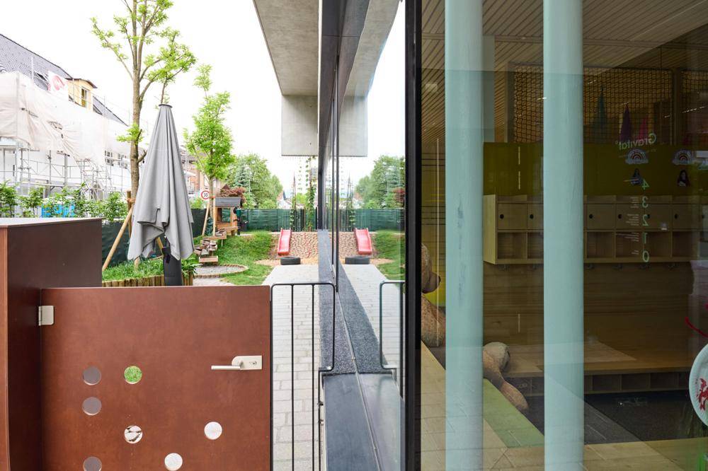 Außenbereich mit Spielplatz und Rutschen, umgeben von Grünflächen, sichtbar durch eine moderne Glasfassade.