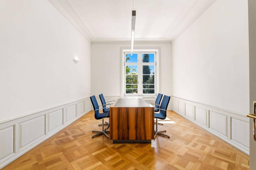 Heller Büroraum mit Parkettboden, hohen Decken und einem großen Fenster mit Blick ins Grüne.