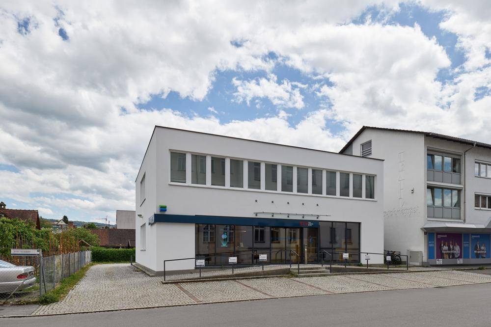 Modernes Geschäftsgebäude mit weißer Fassade, großen Fenstern und einem gepflasterten Parkplatz davor.