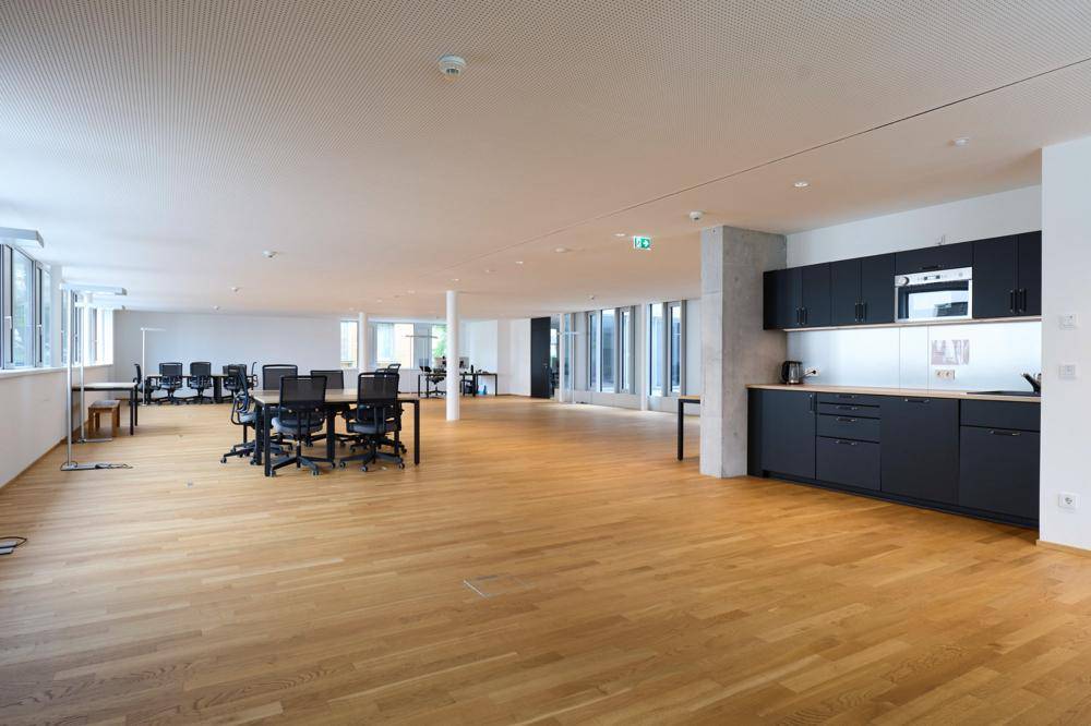 Offener Bürobereich mit integrierter moderner Küchenzeile, Holzboden und vielen Fenstern.
