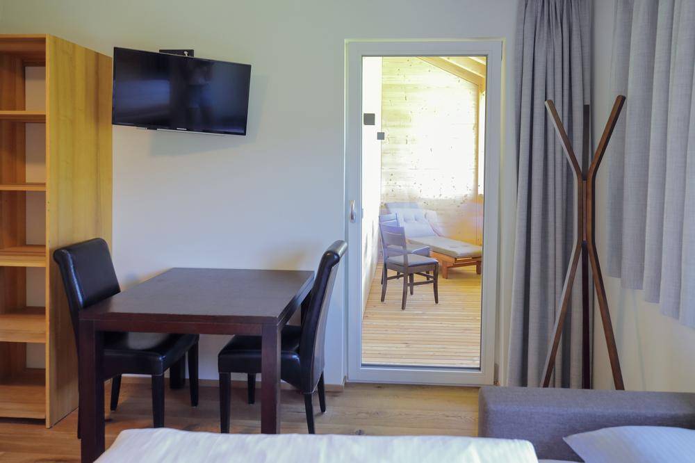 Schlafzimmer mit Esstisch, Fernseher und Zugang zu einem überdachten Balkon mit Holzverkleidung.