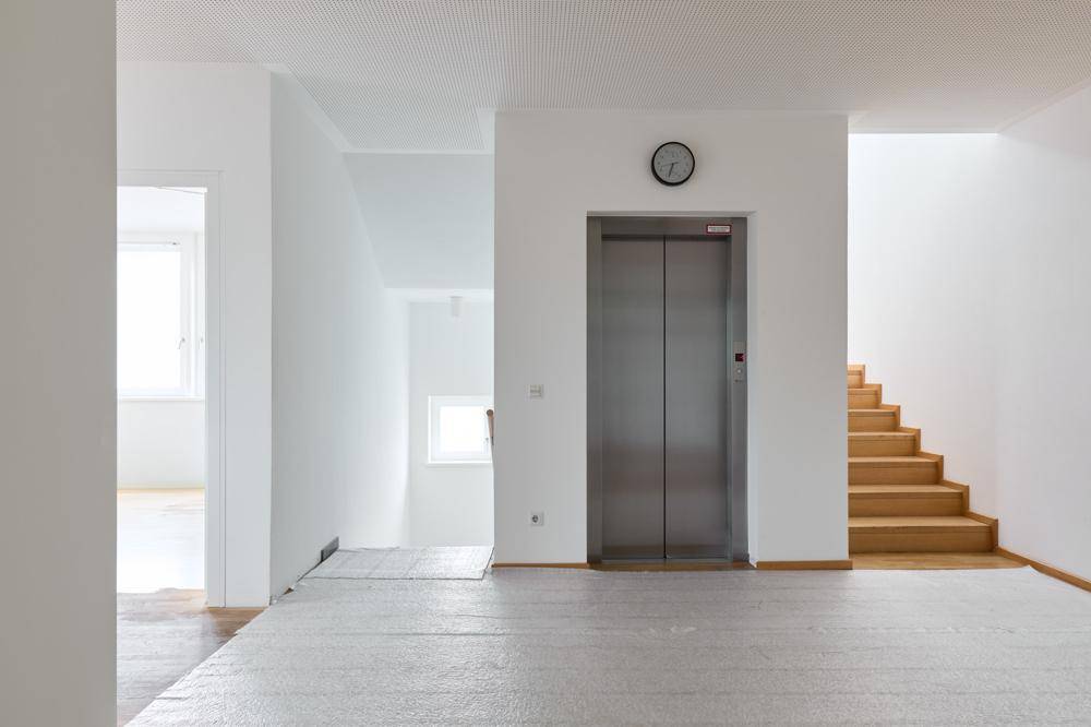 Heller Flur mit Aufzug, Holztreppe und einem kleinen Fenster, das Tageslicht spendet.