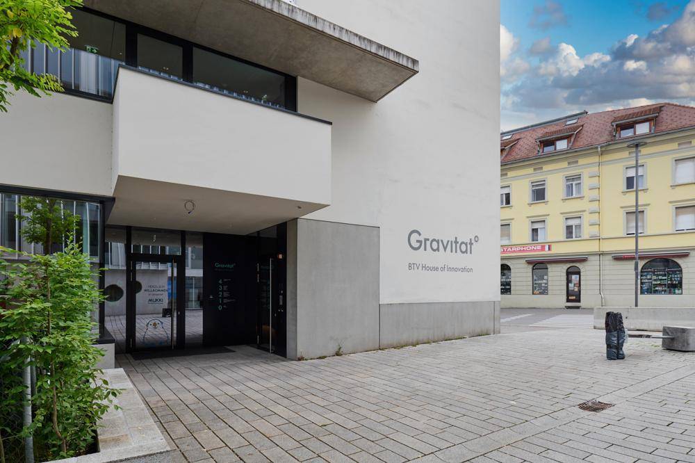 Eingangsbereich des "Gravitat" Gebäudes mit moderner Architektur, umgeben von städtischer Bebauung und gepflastertem Vorplatz.