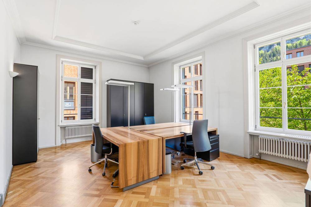Modern eingerichtetes Büro mit Parkettboden, großen Fenstern und Blick auf die umliegenden Gebäude.