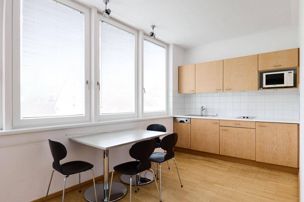 Kleine Küche mit Holzeinbauschränken, Spüle, Mikrowelle und einem Esstisch am Fenster.