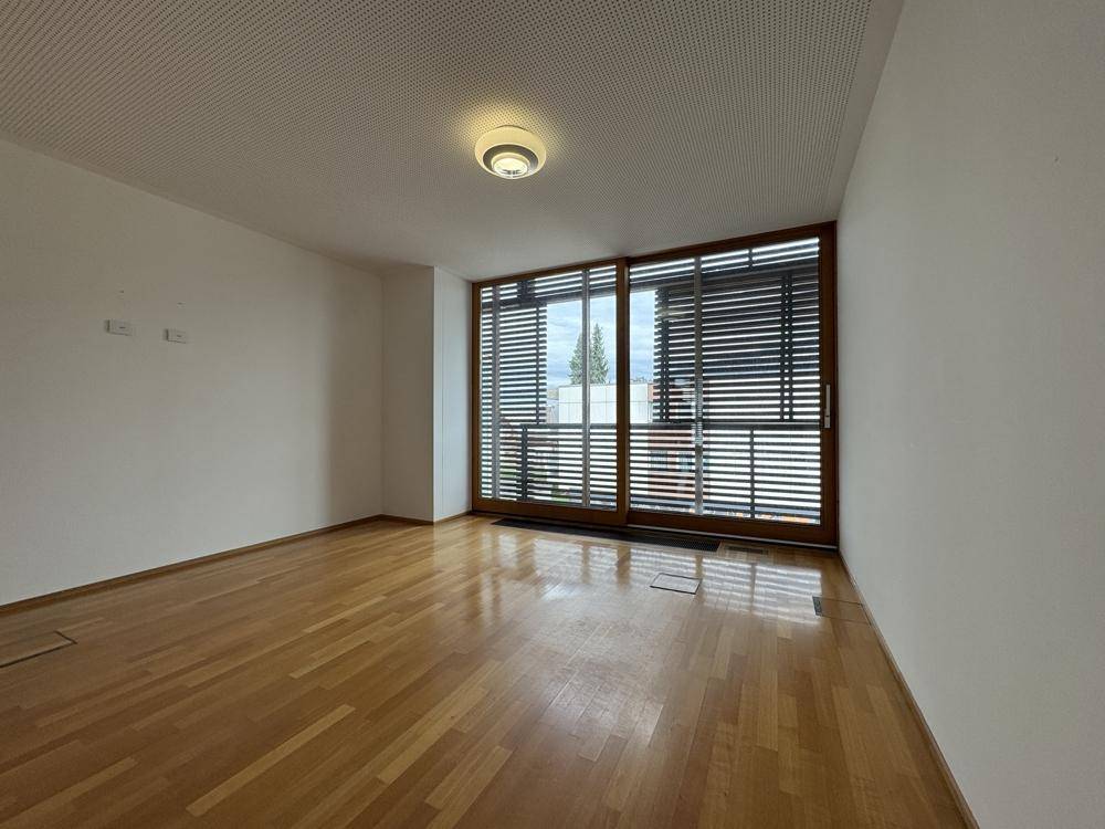 Leerer Raum mit Holzboden und einer großen Fensterfront, die viel Tageslicht hereinlässt.