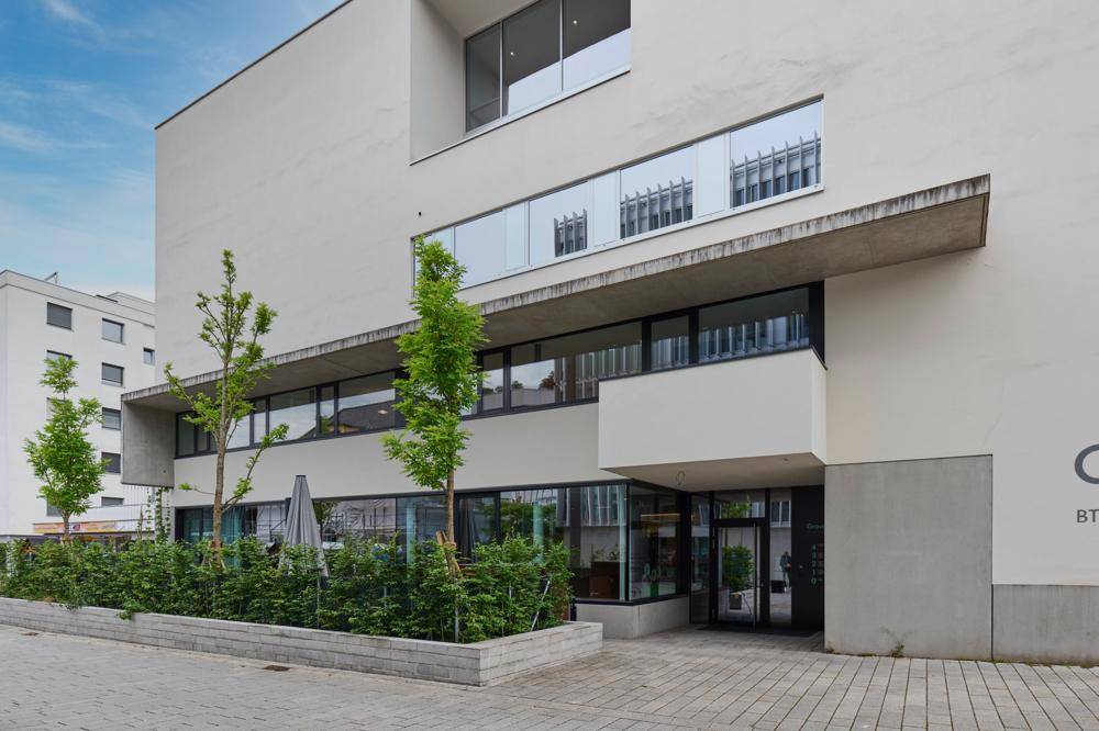 Eingangsbereich des modernen Gebäudes mit gepflegter Bepflanzung und großen Fensterfronten.