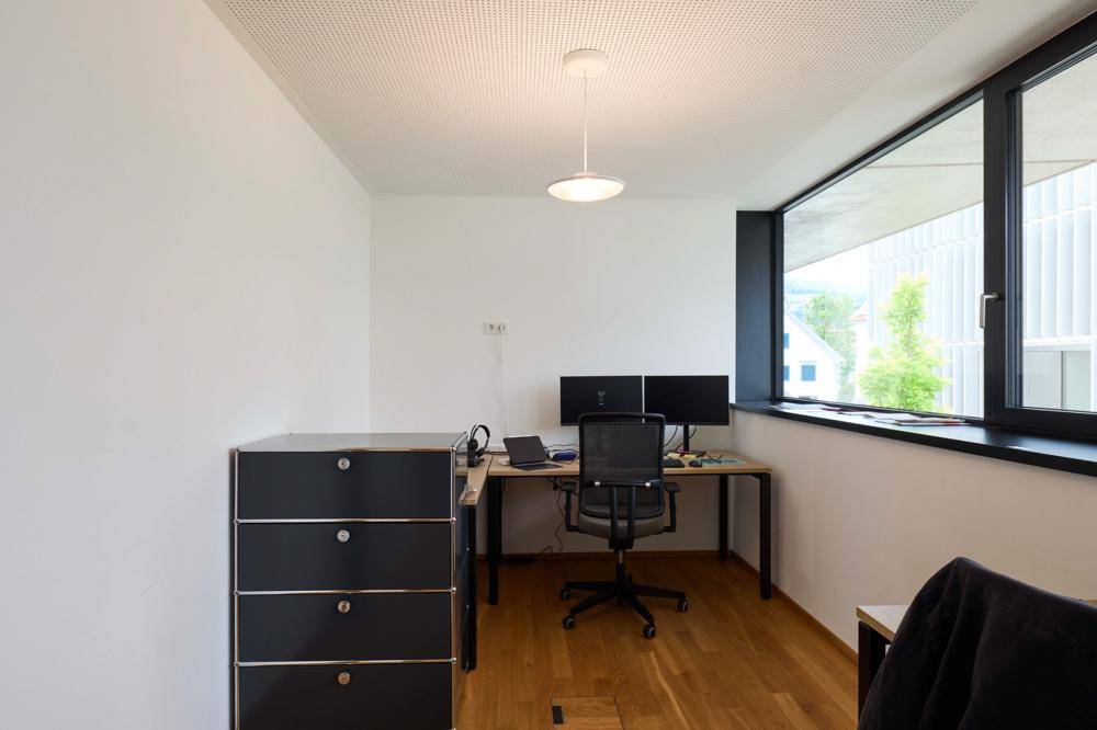 Kleines Büro mit Holzboden, großem Fenster und modernem Schreibtisch mit zwei Monitoren.