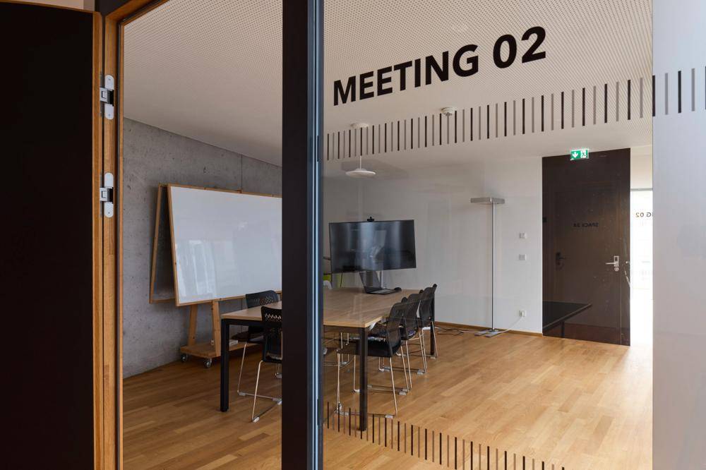 Konferenzraum "Meeting 02" mit Glastür, Holzboden, großem Bildschirm und Whiteboard.
