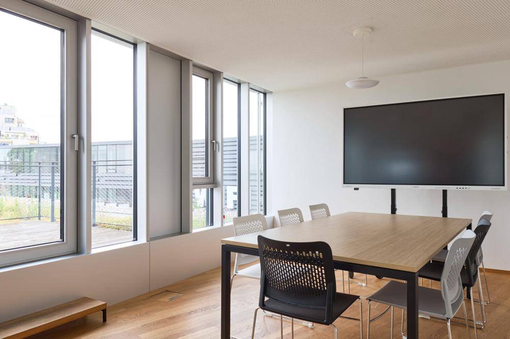 Heller Besprechungsraum mit großem Wandbildschirm, modernem Tisch und Stühlen, bietet viel Tageslicht und Ausblick.