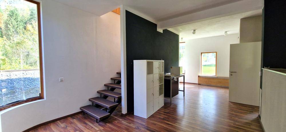 Heller Innenraum mit Holzfußboden, moderner Treppe und großen Fenstern mit Blick ins Grüne.