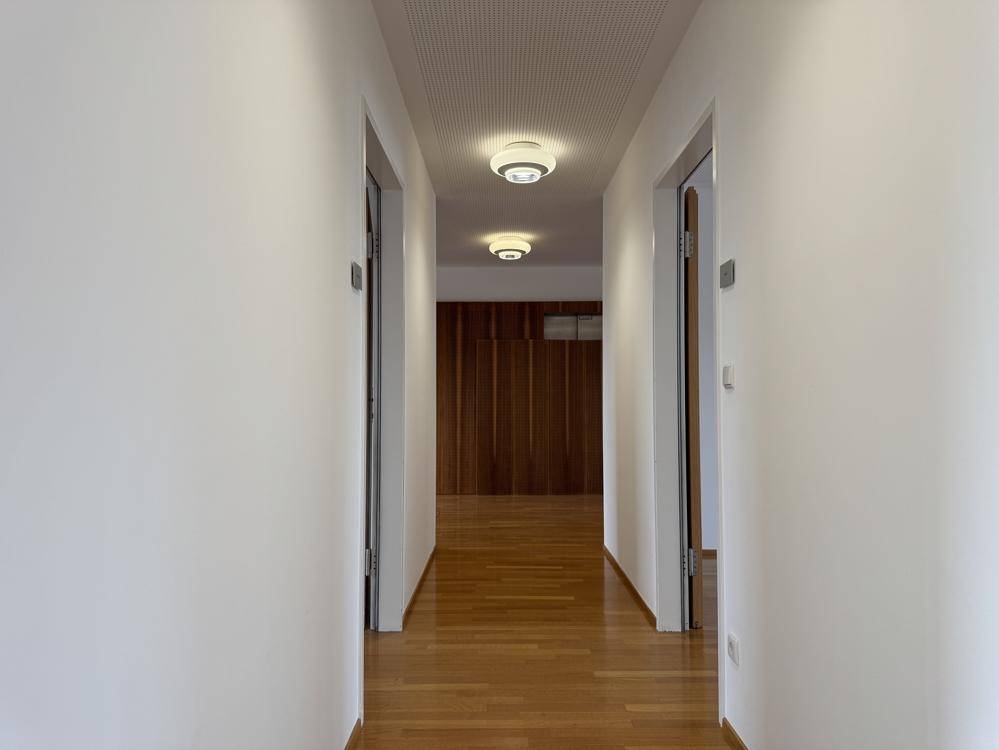 Heller Flur mit Holzparkettboden und Deckenleuchten, der zu einem Bereich mit Holzwand führt.