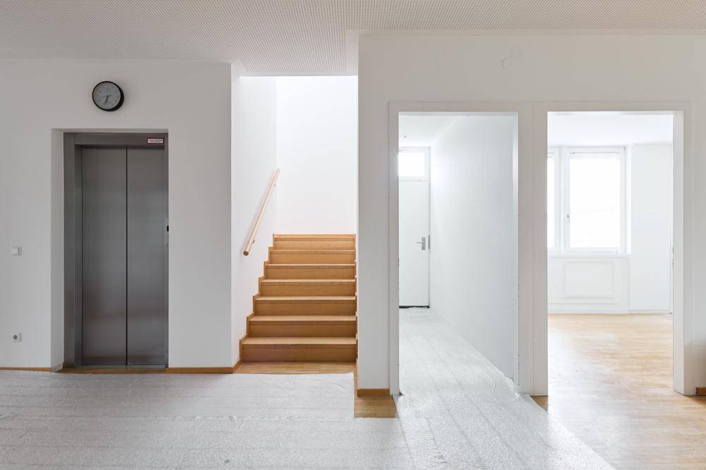 Heller Flur mit Holztreppe, Aufzug und Parkettboden in einem modernen Gebäude.