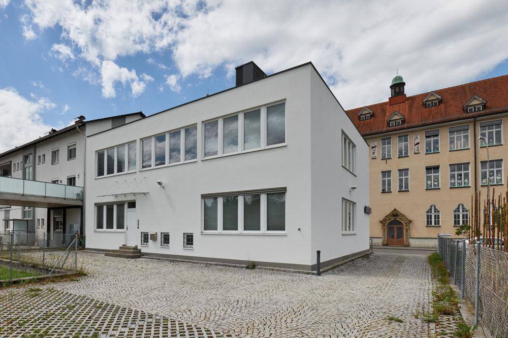 Modernes weißes Gebäude mit großen Fenstern und gepflastertem Vorplatz neben einem älteren Gebäude.