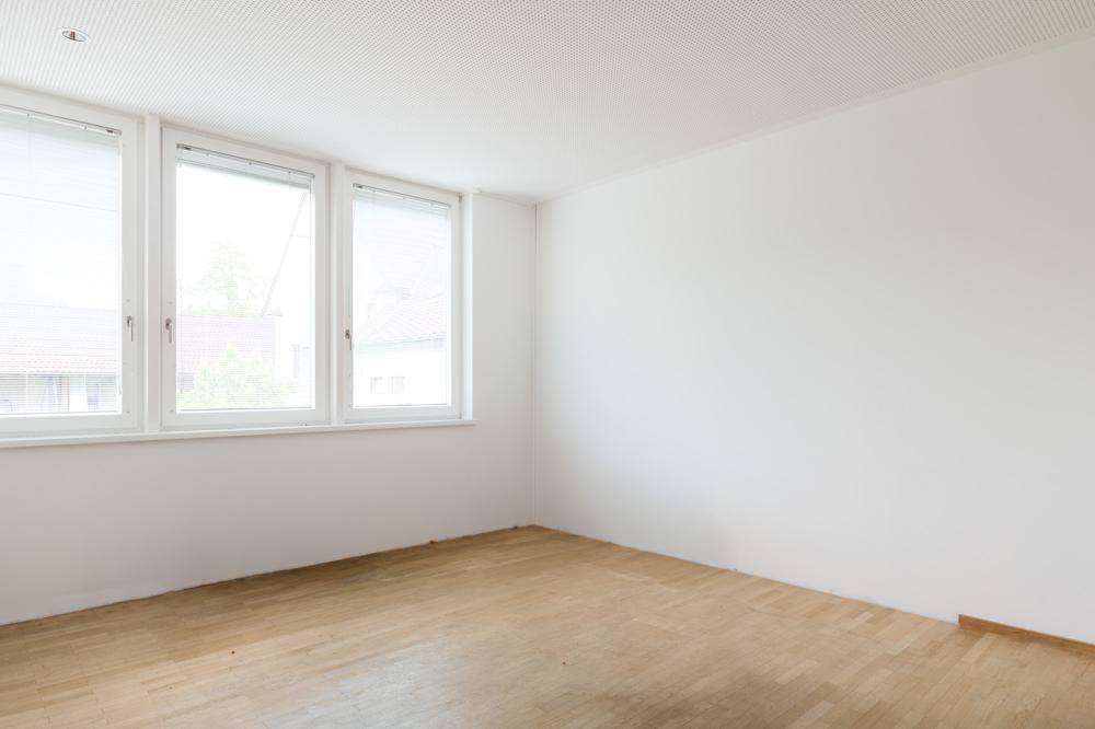 Heller Büroraum mit Parkettboden und drei großen Fenstern, die viel Tageslicht hereinlassen.