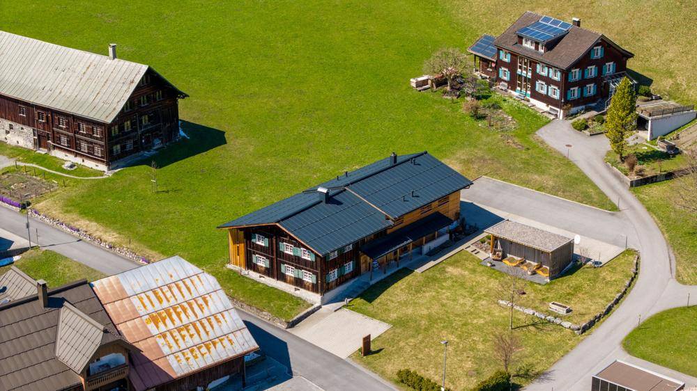 Luftaufnahme von mehreren Häusern in einer grünen, ländlichen Umgebung mit Bergen im Hintergrund.
