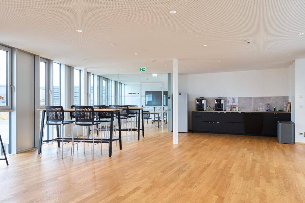 Großer, heller Bürobereich mit hohen Tischen, einer Kaffeestation und großen Fenstern, ideal für Pausen oder informelle Treffen.