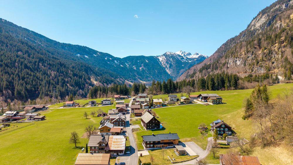 Weitläufige Landschaftsaufnahme eines Dorfes in einem Tal, umgeben von grünen Wiesen und bewaldeten Bergen.