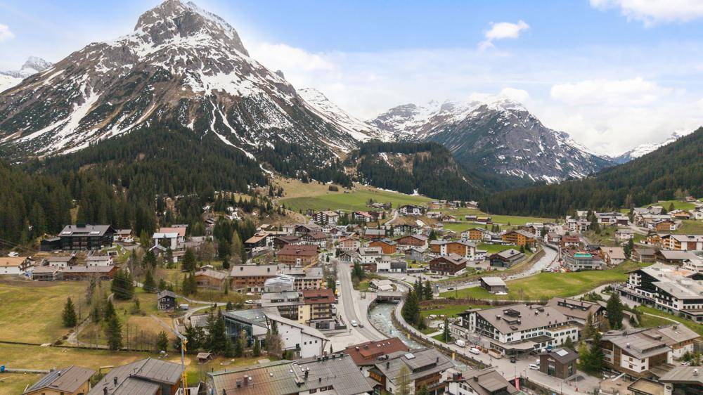 Luftaufnahme eines malerischen Dorfes in einem Tal, umgeben von schneebedeckten Bergen unter blauem Himmel.