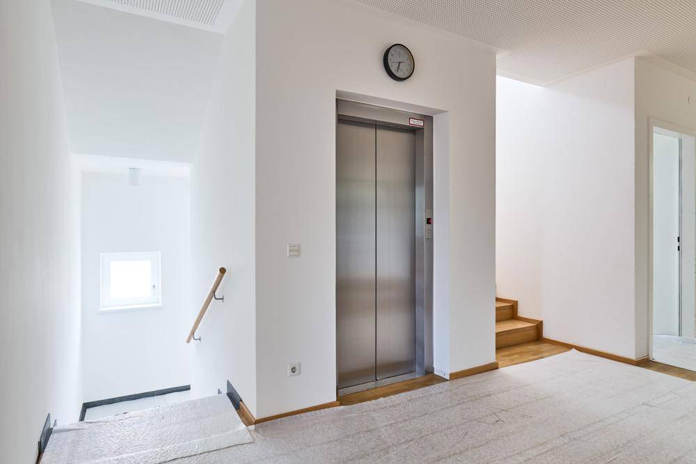 Heller Eingangsbereich mit Aufzug, Holztreppe und einem kleinen Fenster im Treppenhaus.