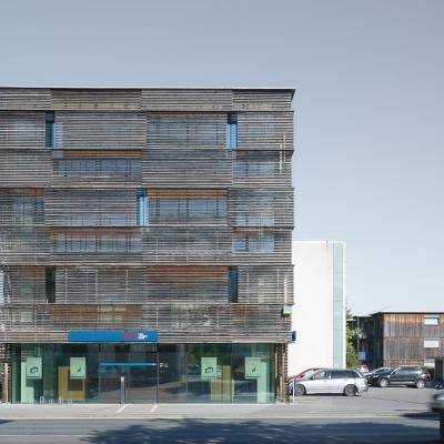 Modernes Gebäude mit Holzlamellenfassade und großen Glasflächen im Erdgeschoss, umgeben von Parkplätzen.