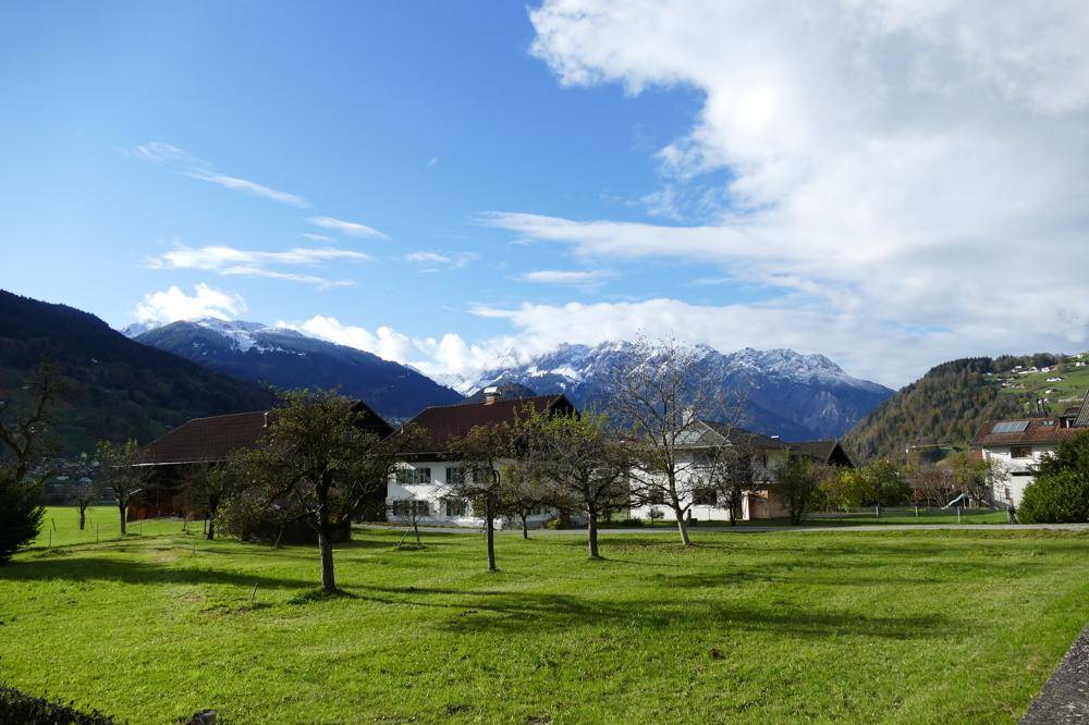 Blick auf eine ländliche Siedlung mit traditionellen Häusern und schneebedeckten Bergen im Hintergrund unter blauem Himmel.