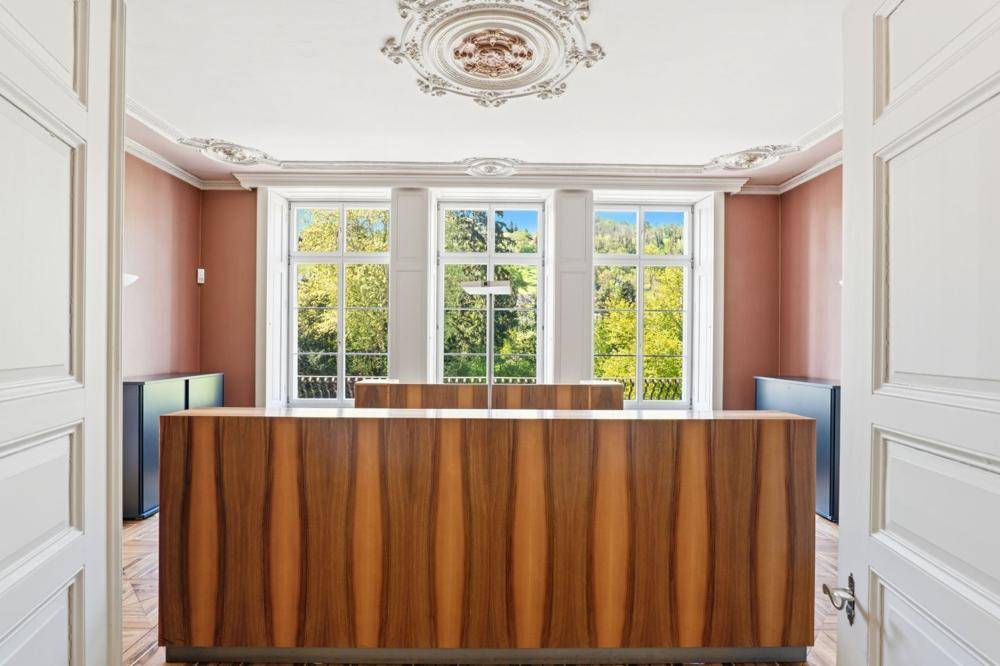 Empfangsbereich mit großer Holztheke, hohen Fenstern und dekorativer Stuckdecke, bietet Blick ins Grüne.