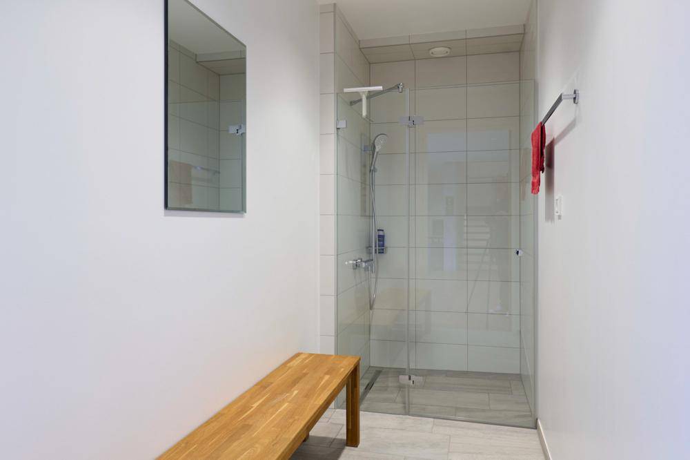 Moderne Duschkabine mit Glaswand, Fliesenboden und einer Holzbank in einem hellen Raum.