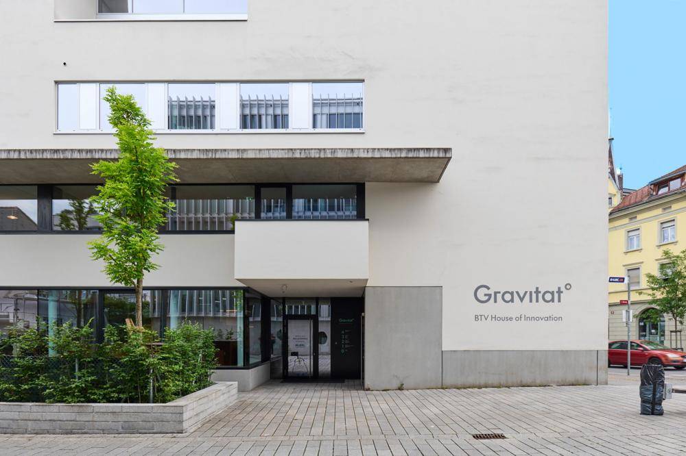 Eingangsbereich des modernen "Gravitat" Gebäudes mit klaren Linien, großen Fenstern und gepflegter Bepflanzung.
