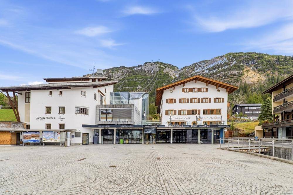 Zwei Gebäude im alpinen Stil mit Geschäften im Erdgeschoss und Bergen im Hintergrund unter klarem Himmel.