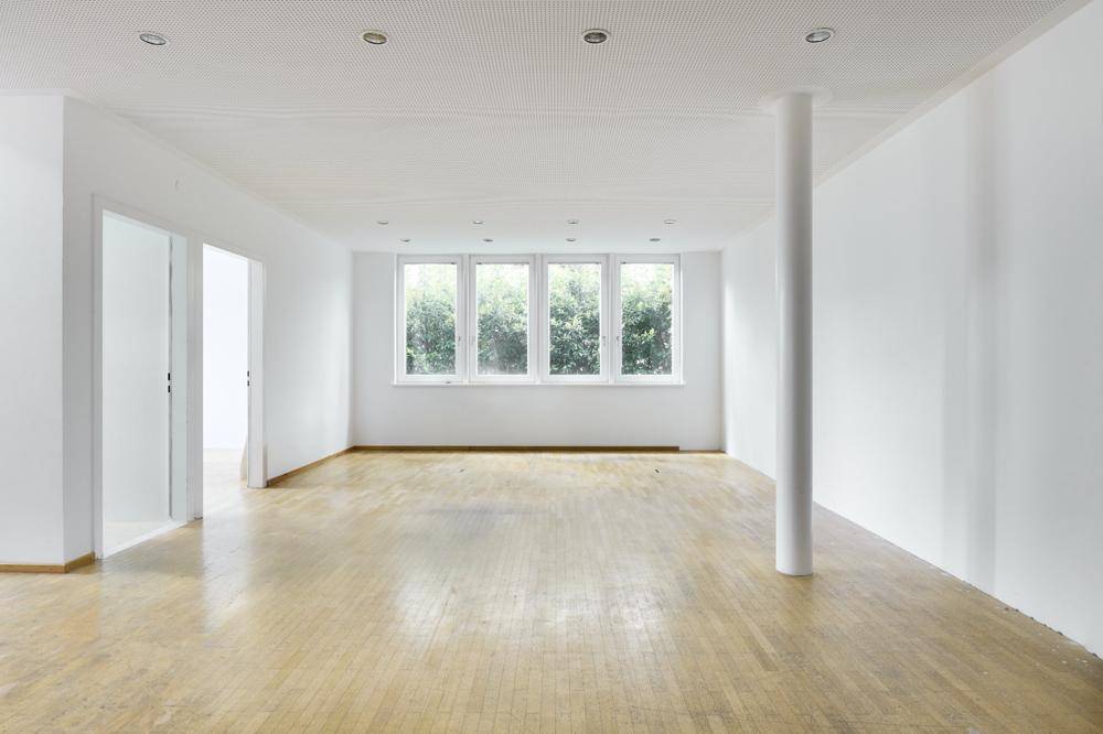 Geräumiger, leerer Büroraum mit hellen Wänden, Parkettboden und großen Fenstern mit Grünblick.