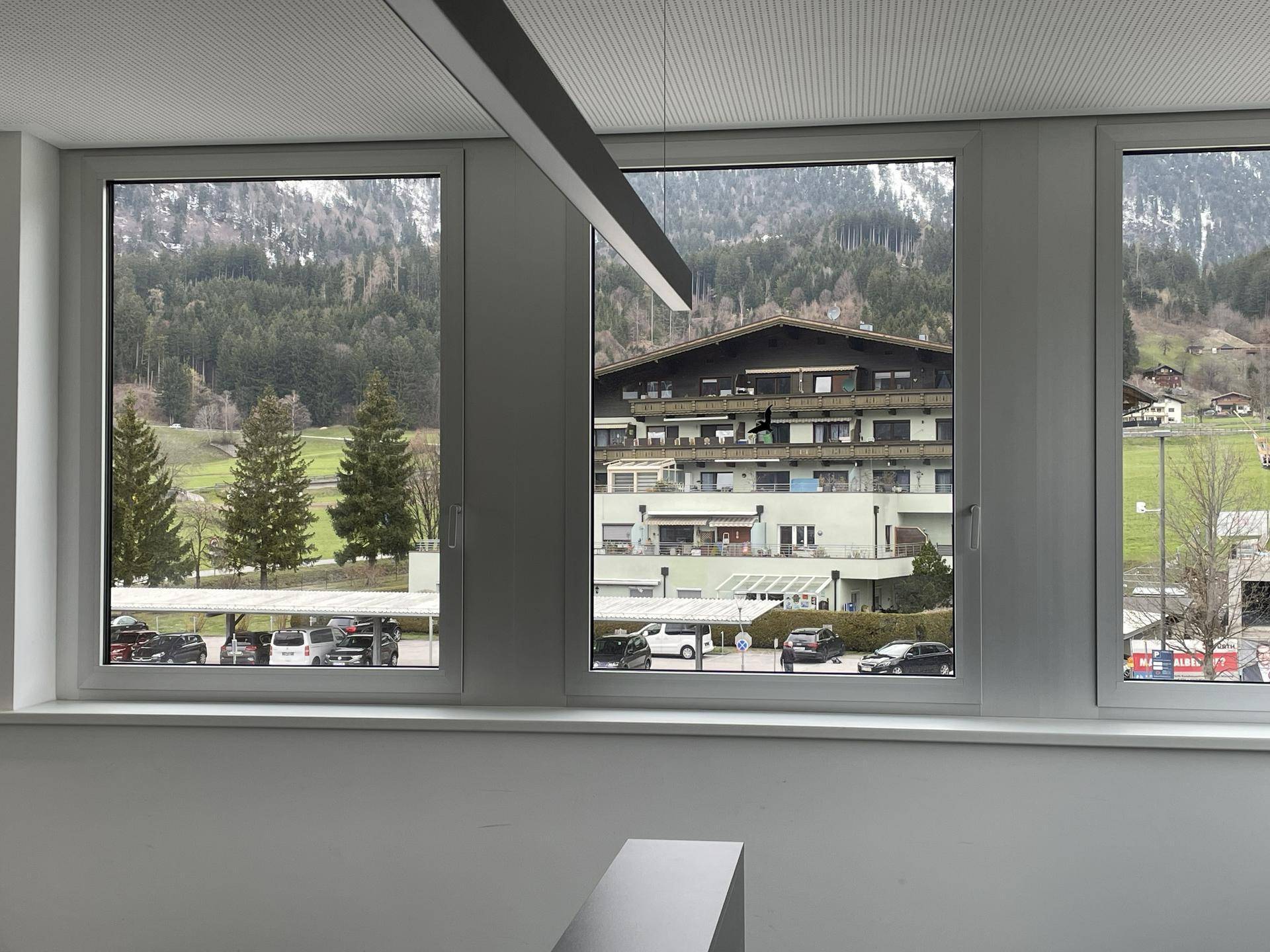 Fensterblick aus einem Büro auf ein gegenüberliegendes Gebäude, Parkplätze und die umliegende Berglandschaft.