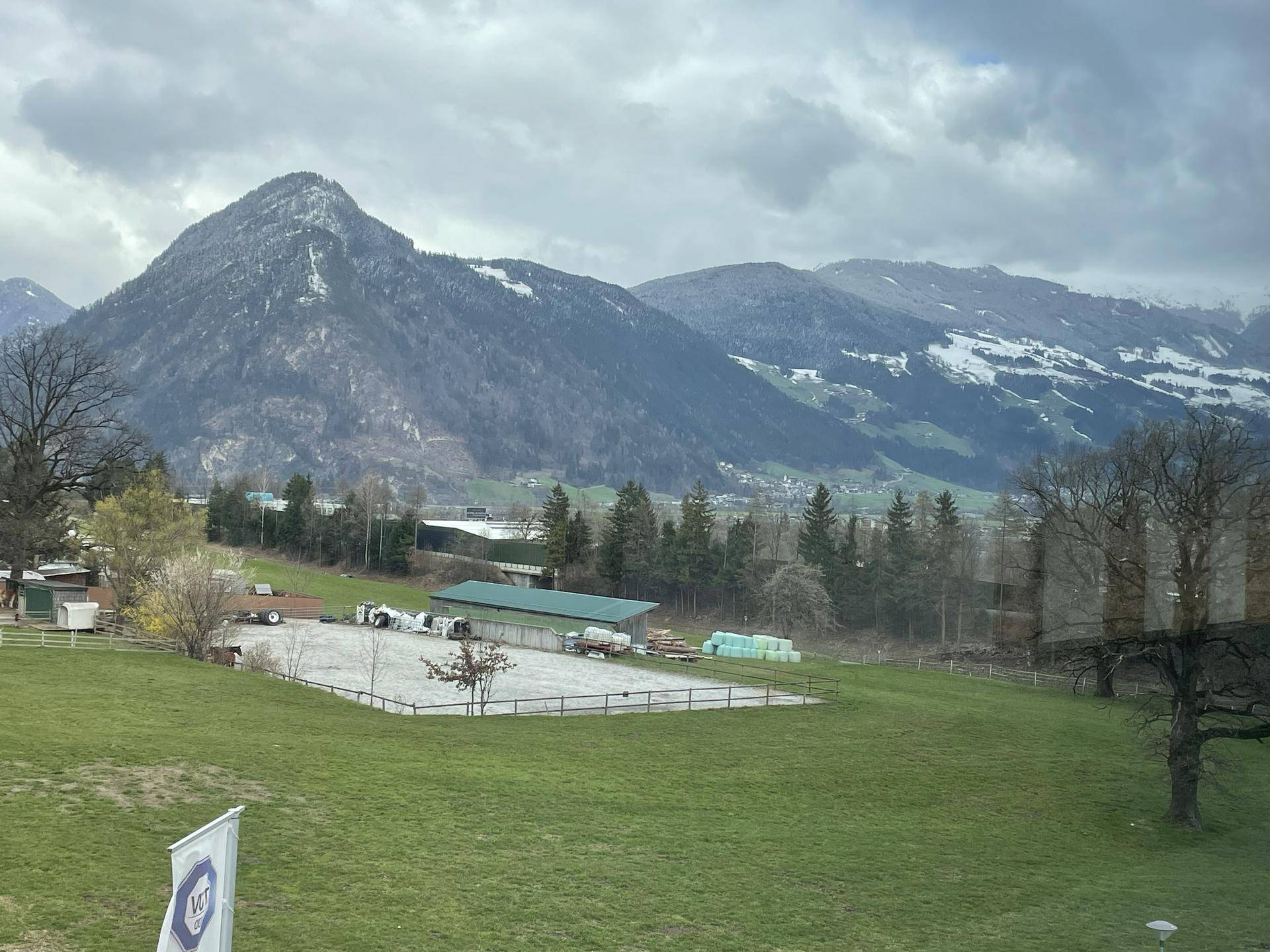 Panoramablick aus dem Fenster auf eine weite grüne Landschaft mit majestätischen Bergen und einem Reitplatz.