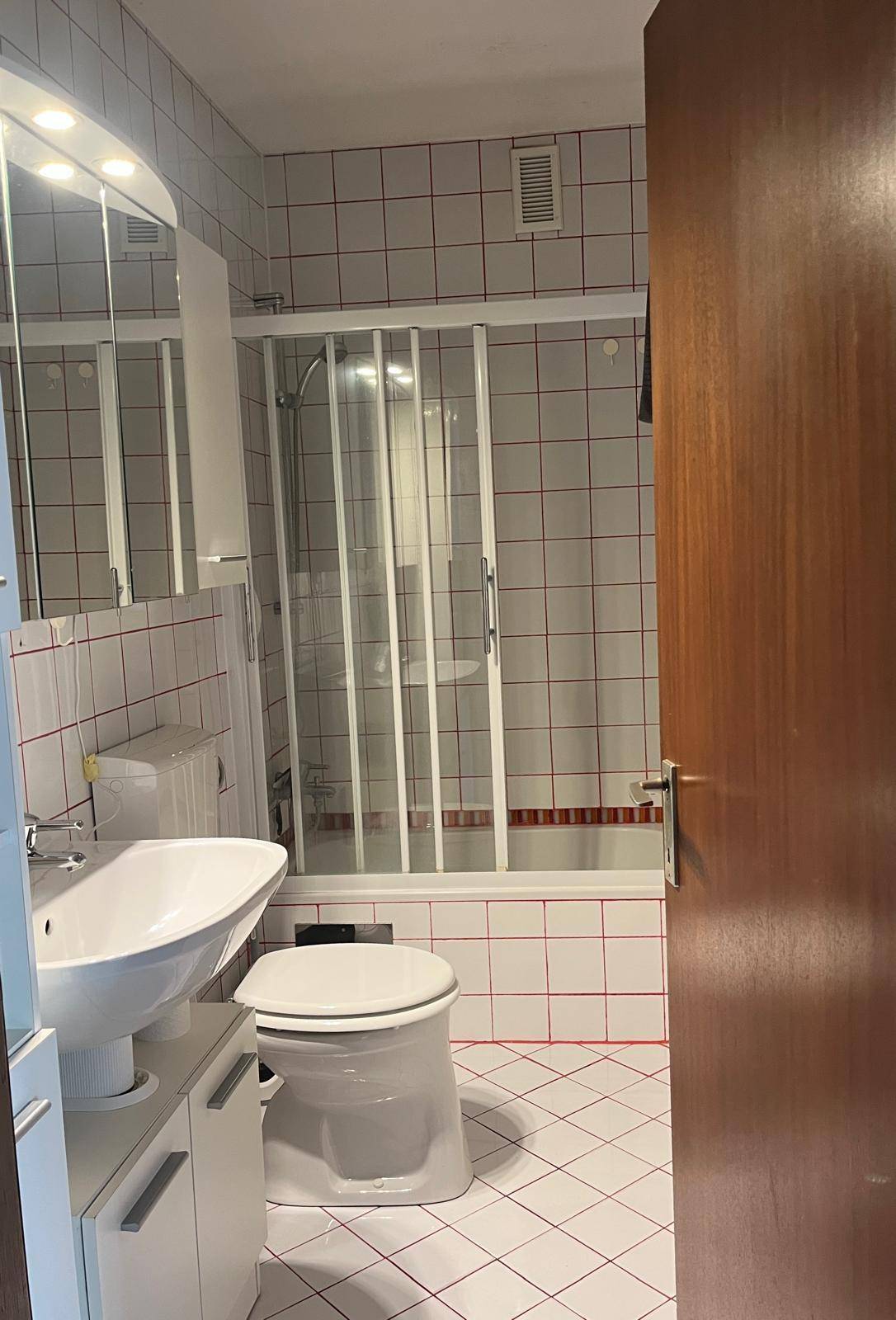 Badezimmer mit weißen Fliesen, rotem Fugenmuster, Badewanne mit Duschabtrennung und Waschbecken.