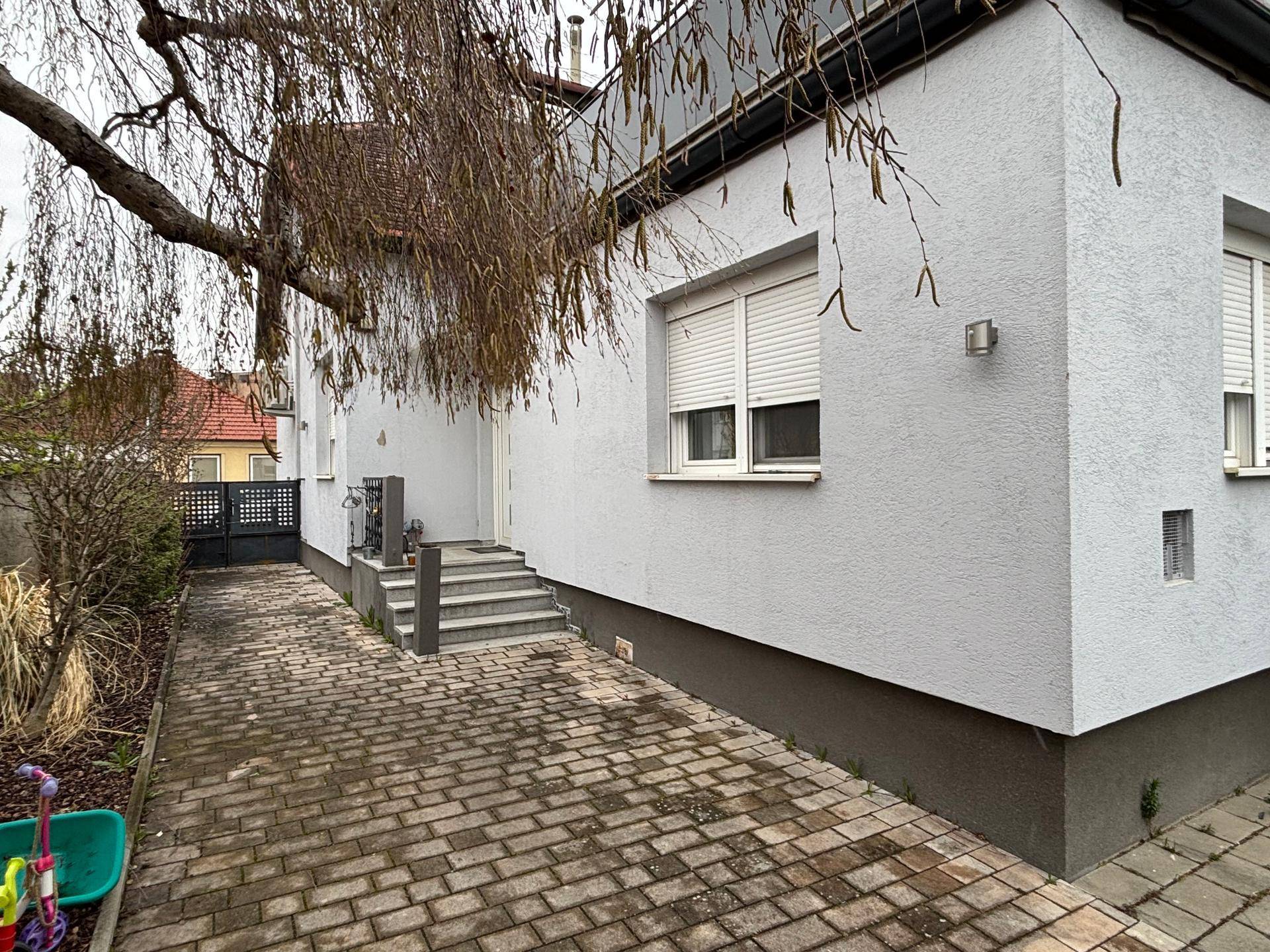 Seitenansicht des Hauses mit gepflastertem Zugang und einem Baum im Vordergrund.