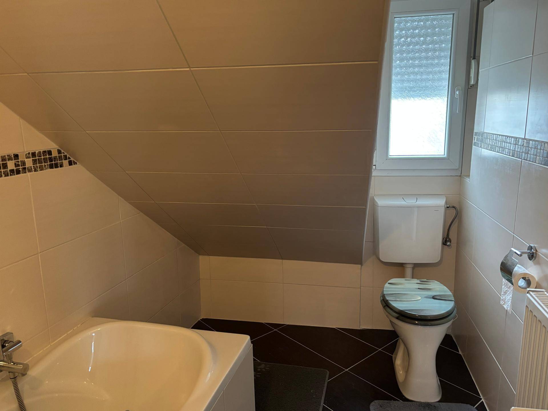 Badezimmer im Dachgeschoss mit Badewanne, WC und Schrägdach, hell gefliest.