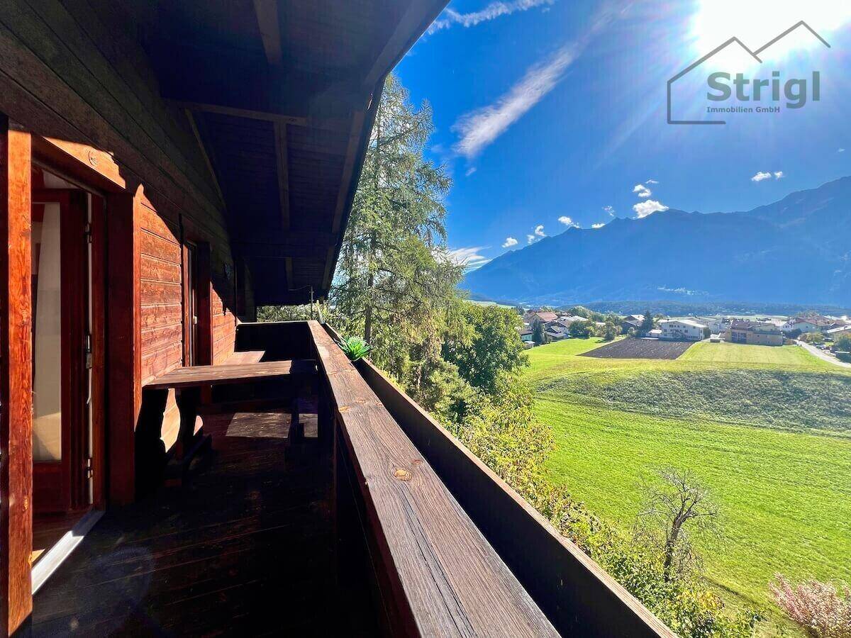 Geräumiger Holzbalkon mit Sitzgelegenheiten und weitem Blick über grüne Felder und Berge.