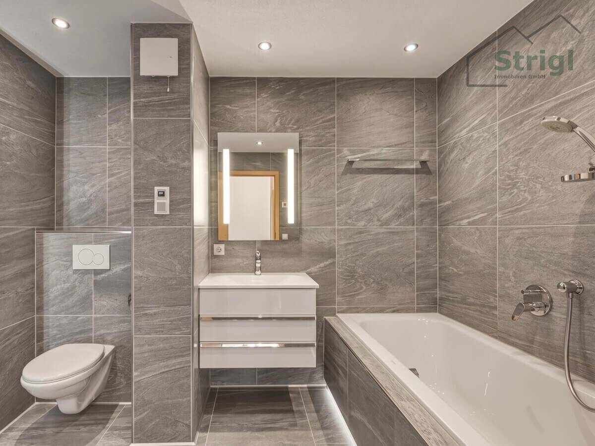 Modernes Badezimmer mit grauen Fliesen, Badewanne und wandhängendem WC.