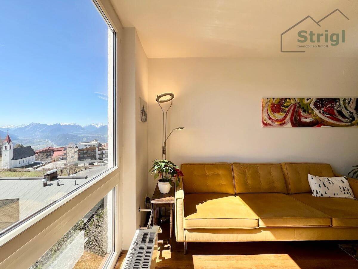 Helles Wohnzimmer mit großem Fenster, das einen weiten Blick auf die Berge und das Dorf bietet.