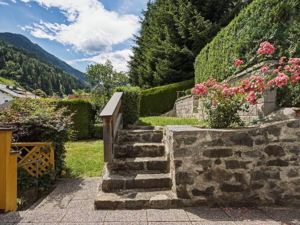 Gepflegter Gartenbereich mit Steintreppe, Hecken und blühenden Sträuchern vor Bergkulisse.