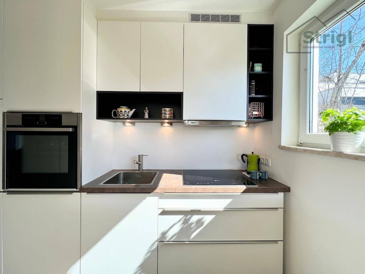 Moderne Küche mit weißen Schränken, integriertem Backofen und Induktionskochfeld, hell durch ein Fenster.
