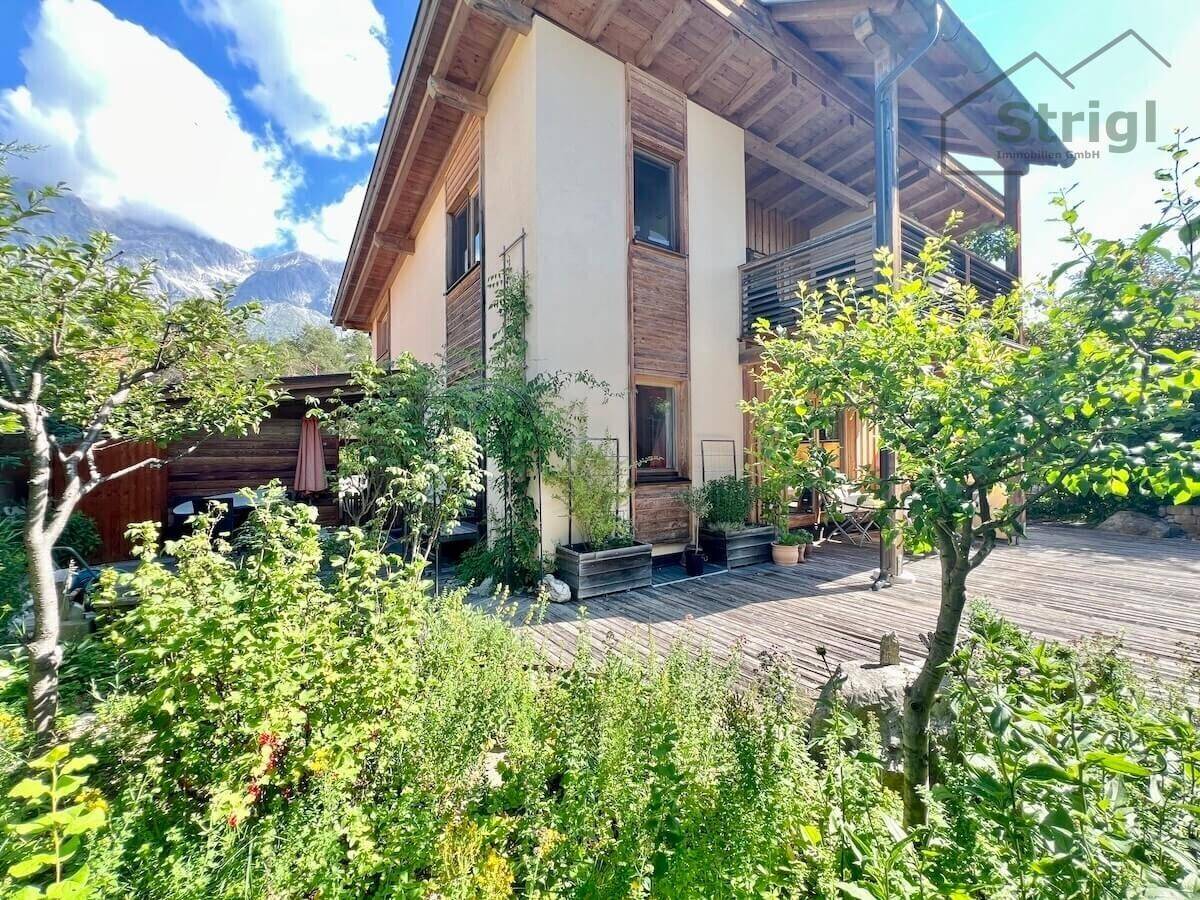 Modernes Wohnhaus mit Holzfassade, großer Holzterrasse und üppigem Garten vor Bergpanorama.