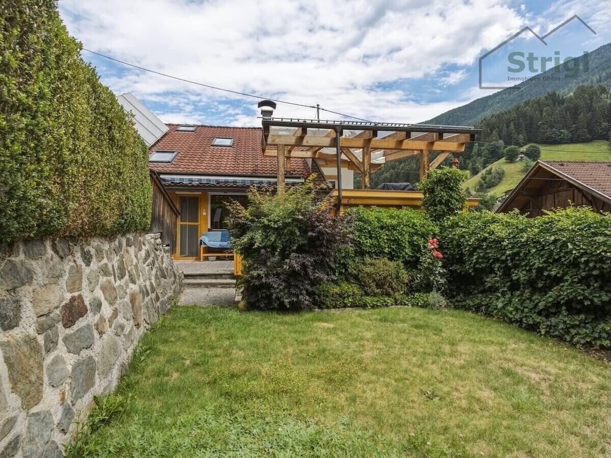 Hinterer Gartenbereich mit Rasenfläche, hohen Hecken und einer Terrasse mit Bergblick.