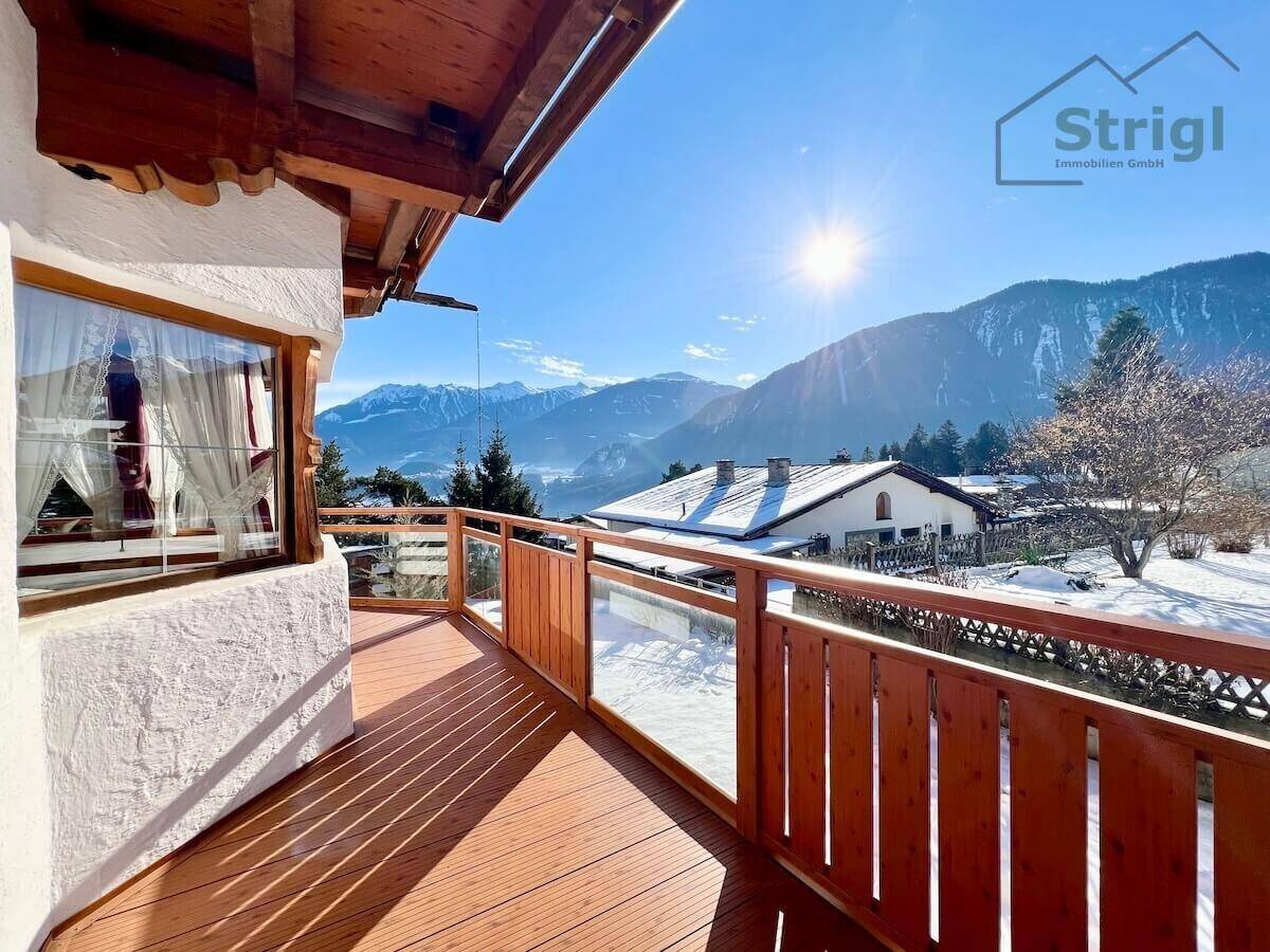 Sonniger Balkon mit Holzboden und Glasgeländer, bietet einen weiten Blick auf die verschneiten Berge.